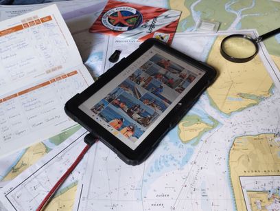 A rugged tablet showing a grid of shared sailing photos, resting on printed nautical charts and a dive logbook